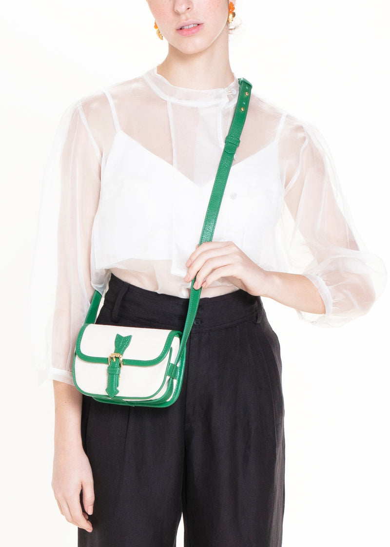 Bolsa de canvas cru com acabamento em couro verde e fivela dourada, versátil para ser usada tanto como crossbody com alça longa quanto como pochete ajustada na cintura.