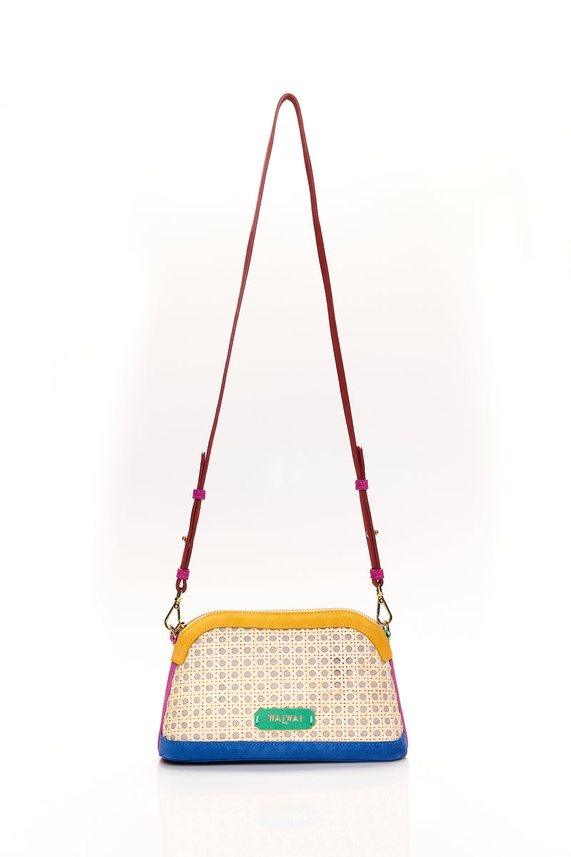 Bolsa estruturada com corpo em palha tramada natural, acabamentos em camurça colorida — amarelo, azul e pink — e alça tiracolo regulável em couro vermelho. Detalhe de plaquinha verde com logo na frente.