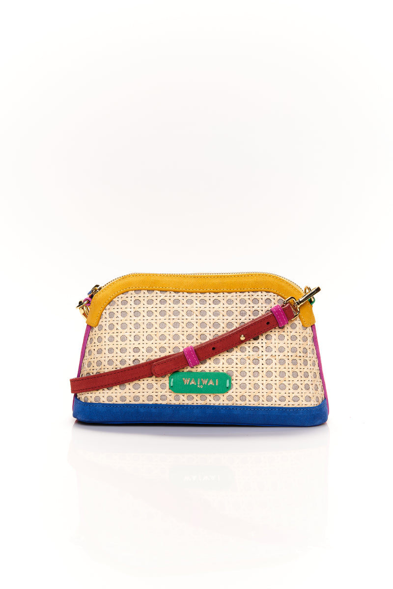 Bolsa estruturada com corpo em palha tramada natural, acabamentos em camurça colorida — amarelo, azul e pink — e alça tiracolo regulável em couro vermelho. Detalhe de plaquinha verde com logo na frente.