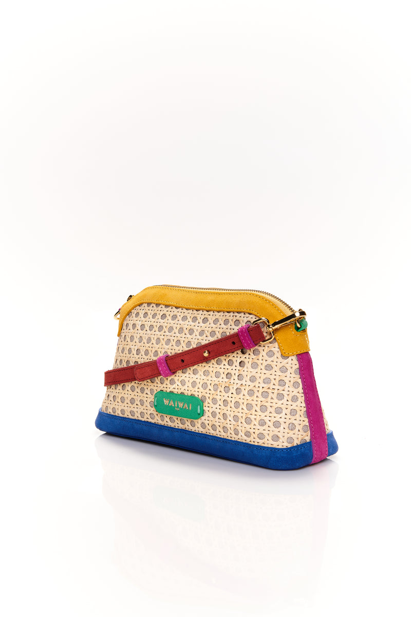 Bolsa estruturada com corpo em palha tramada natural, acabamentos em camurça colorida — amarelo, azul e pink — e alça tiracolo regulável em couro vermelho. Detalhe de plaquinha verde com logo na frente.
