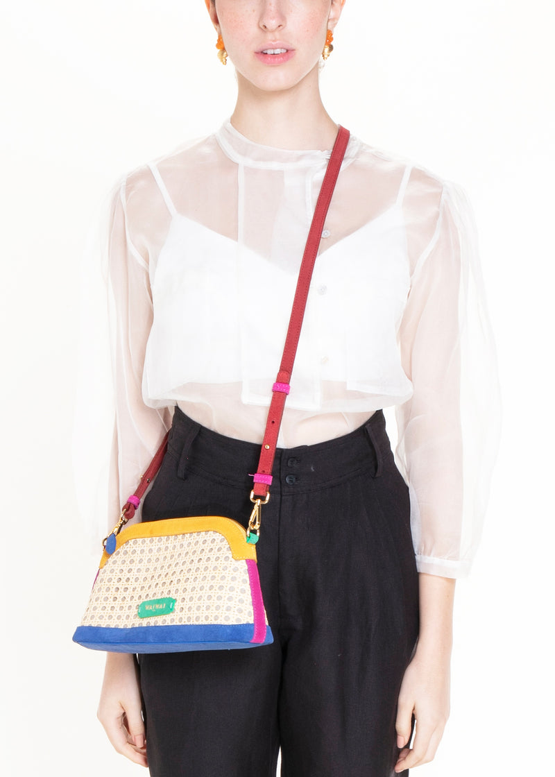 Bolsa estruturada com corpo em palha tramada natural, acabamentos em camurça colorida — amarelo, azul e pink — e alça tiracolo regulável em couro vermelho. Detalhe de plaquinha verde com logo na frente.