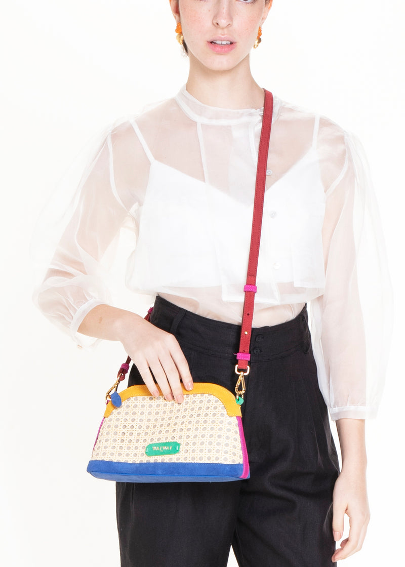 Bolsa estruturada com corpo em palha tramada natural, acabamentos em camurça colorida — amarelo, azul e pink — e alça tiracolo regulável em couro vermelho. Detalhe de plaquinha verde com logo na frente.