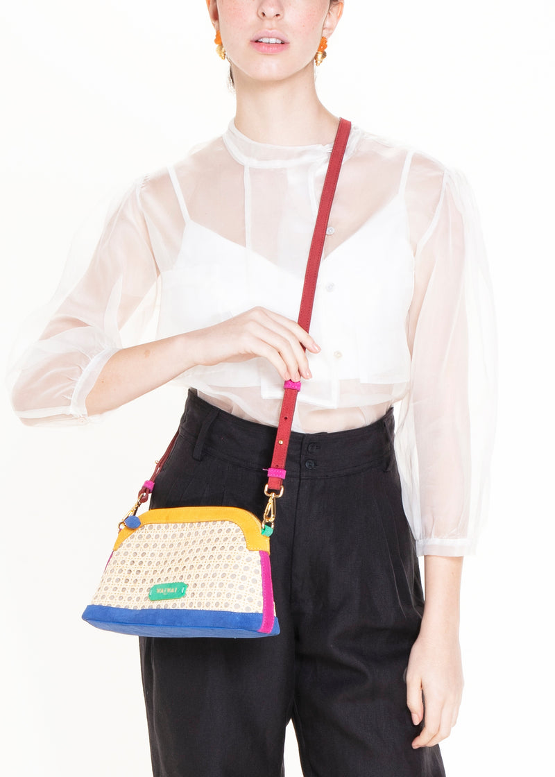 Bolsa estruturada com corpo em palha tramada natural, acabamentos em camurça colorida — amarelo, azul e pink — e alça tiracolo regulável em couro vermelho. Detalhe de plaquinha verde com logo na frente.