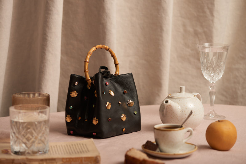 Bolsa em couro preto com aplicações de conchas douradas e cristais coloridos, alça rígida em bambu.