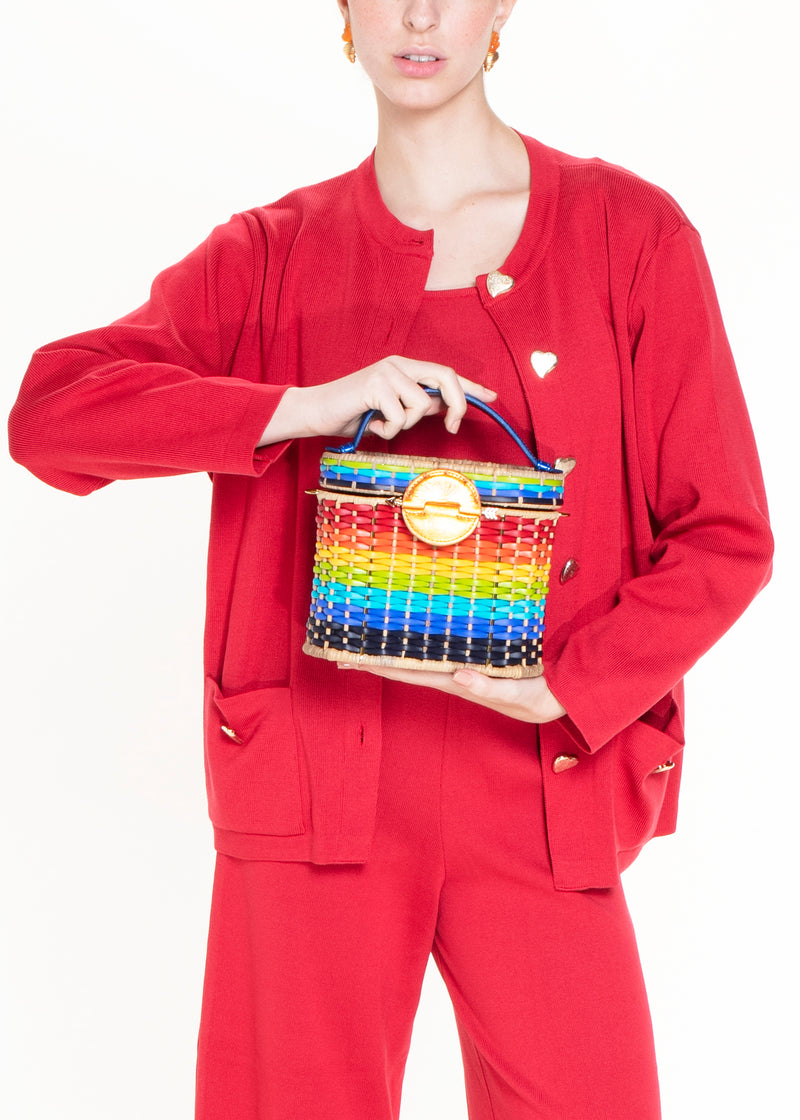 Bolsa em palha com trama degradê rainbow estruturada, fecho circular em couro laranja com símbolo solar e alça curta em couro azul.