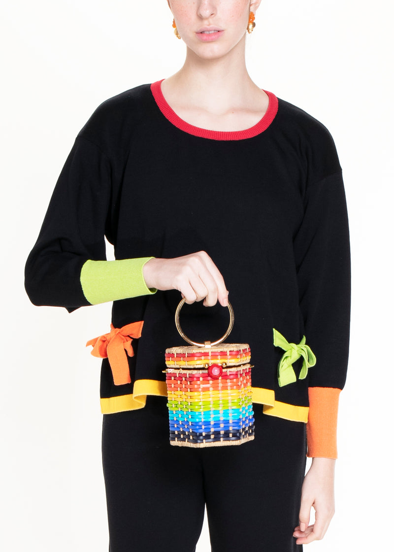 Bolsa estruturada em rattan com trama em degradê de cores do arco-íris, fecho esférico vermelho e alça metálica circular dourada.