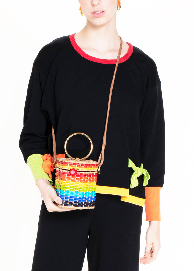 Bolsa estruturada em rattan com trama em degradê de cores do arco-íris, fecho esférico vermelho e alça metálica circular dourada.