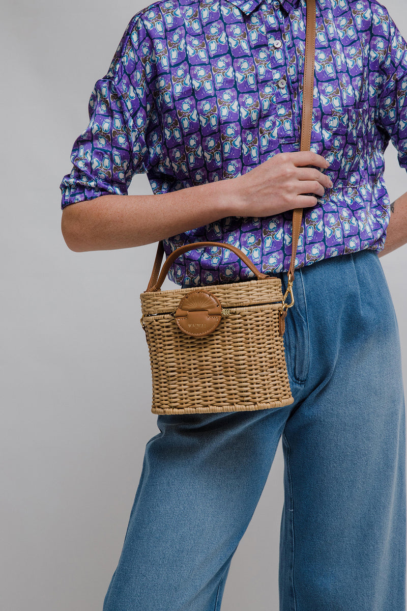 Bolsa de rattan natural com alça curta em couro bege e fecho circular em couro com detalhe em formato de raio de sol dourado.
