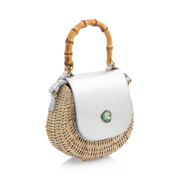 Bolsa em rattan natural estruturada, com tampa em couro prateado metalizado, fecho circular em madrepérola e alça de bambu curvado.