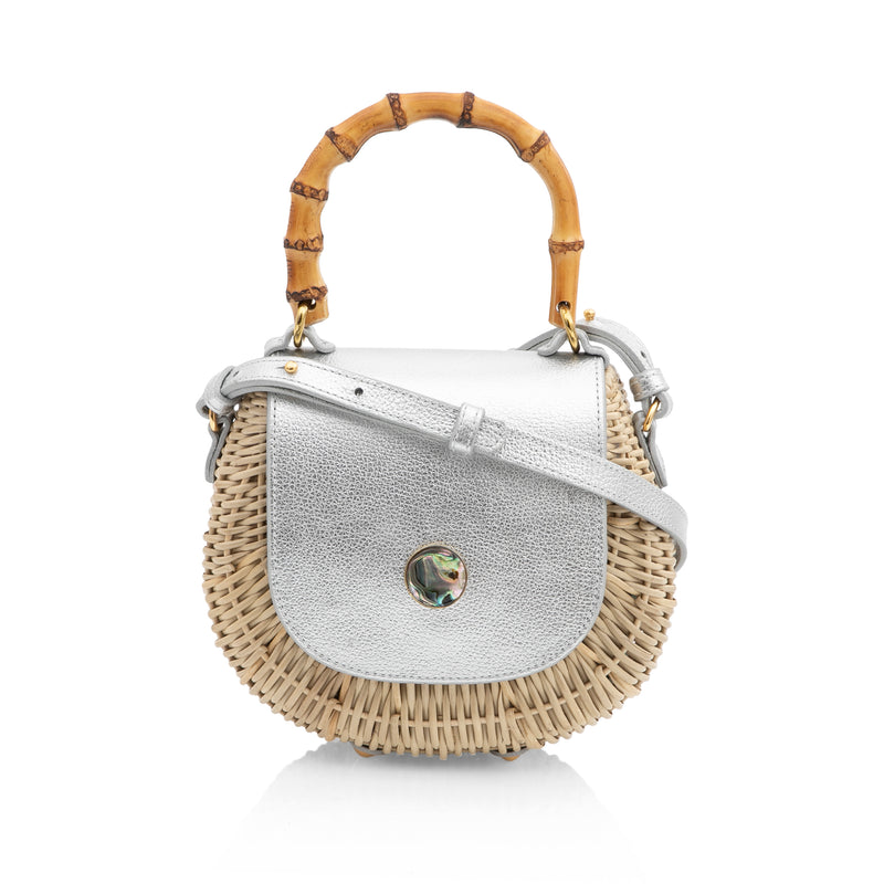 Bolsa artesanal estruturada em rattan natural, com tampa em couro prateado metalizado, fecho circular de madrepérola e alça de bambu curvado.