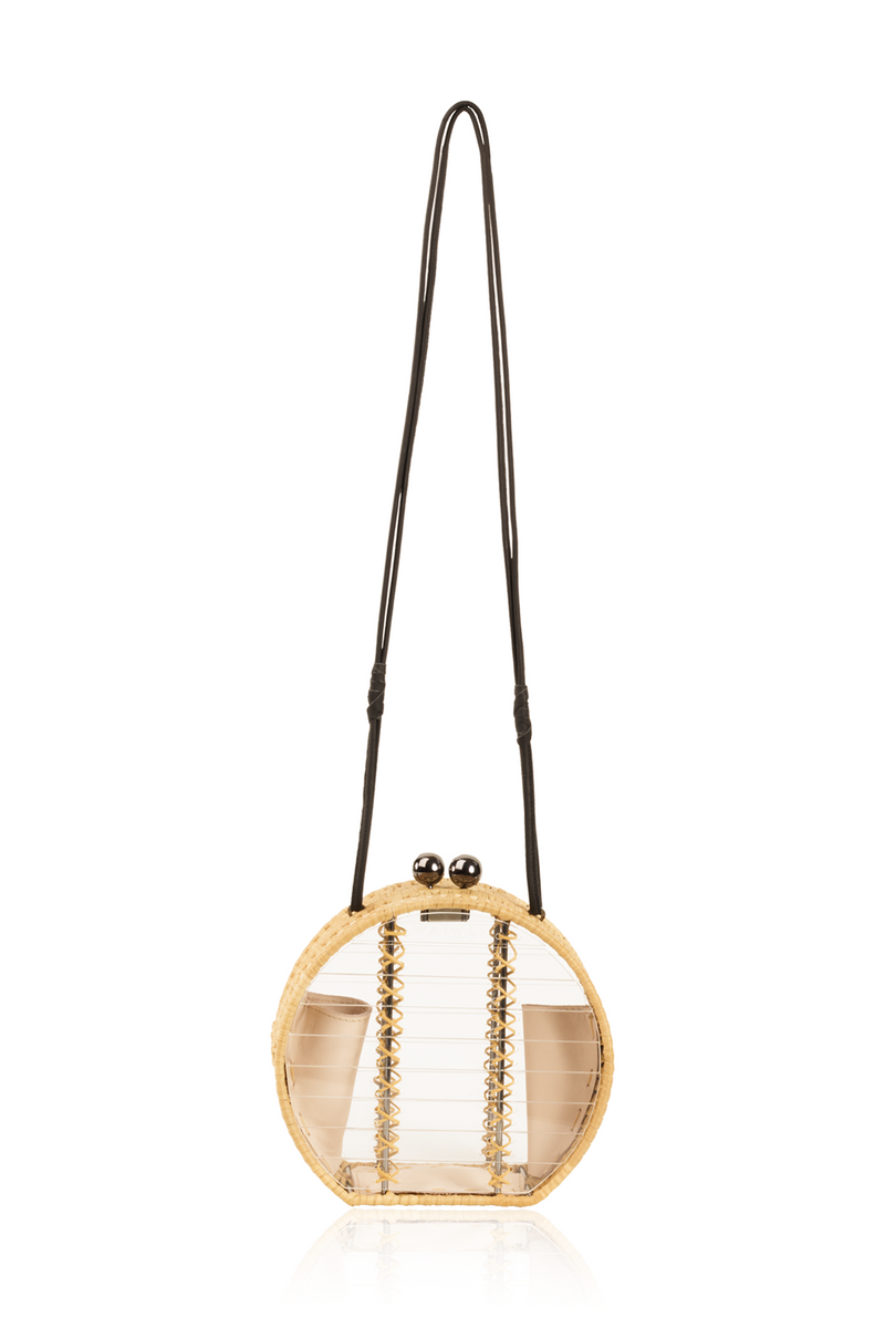 Bolsa redonda em acrílico transparente com laterais em rattan, detalhe de costura em X dourado, alça longa em couro preto e fecho esférico metálico.