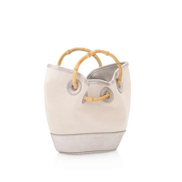 Bolsa bucket em canvas cru, com acabamento em couro prata e alça estruturada de bambu curvado. Detalhes em couro na base e na parte superior.