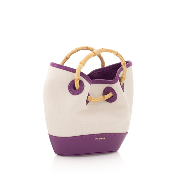 Bolsa bucket em canvas cru, com acabamento em couro roxo e alça estruturada de bambu curvado. Detalhes em couro na base e na parte superior.