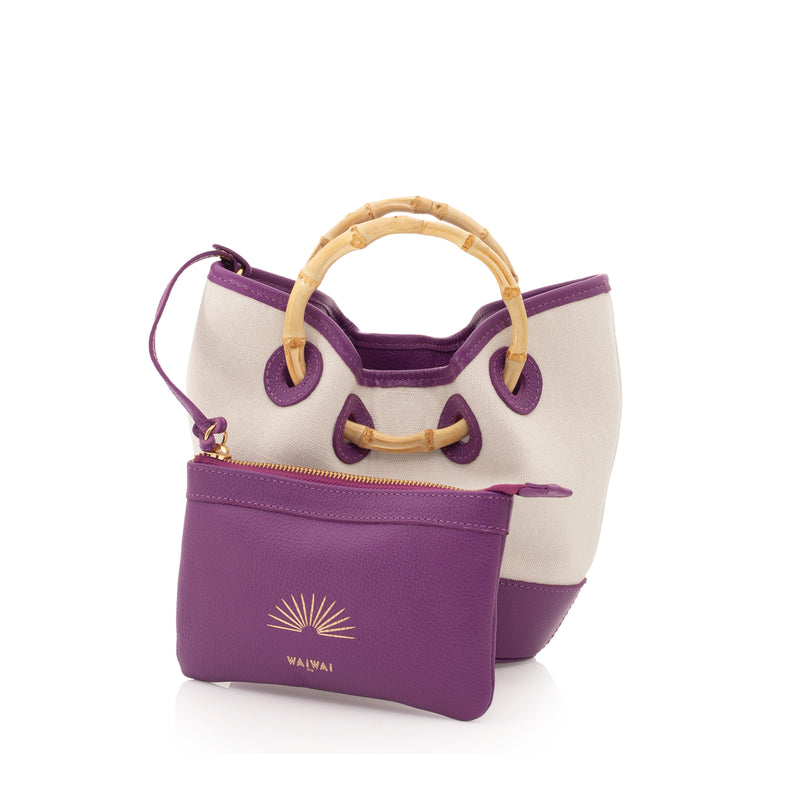 Bolsa bucket estruturada em canvas cru com couro roxo nos acabamentos, alça de bambu curvado e reforço em couro na base.