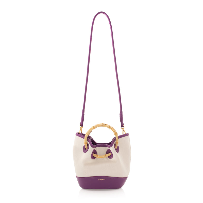 Bolsa bucket em canvas cru com detalhes em couro roxo, alça curta de bambu curvado e acabamento superior e inferior em couro.