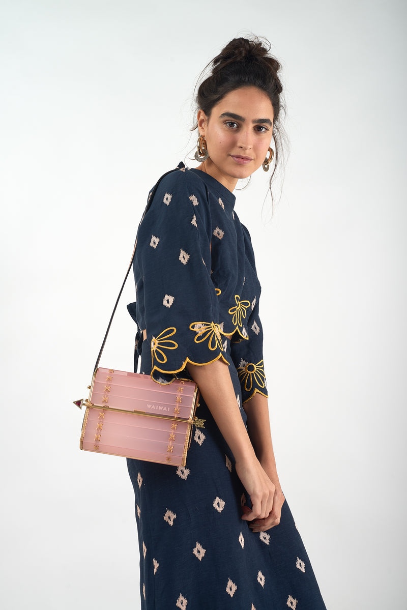 Clutch em acrílico rosa translúcido com laterais em rattan, detalhe de costura em X dourado e fecho em formato de flecha metálica dourada com ponta vermelha.