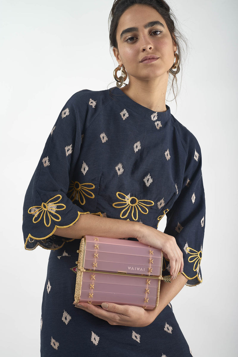 Clutch em acrílico rosa translúcido com laterais em rattan, detalhe de costura em X dourado e fecho em formato de flecha metálica dourada com ponta vermelha.