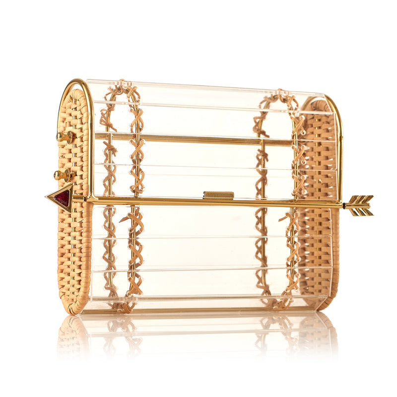 Clutch em acrílico transparente com laterais em rattan, detalhe de costura em X dourado e fecho em formato de flecha metálica dourada com ponta vermelha.