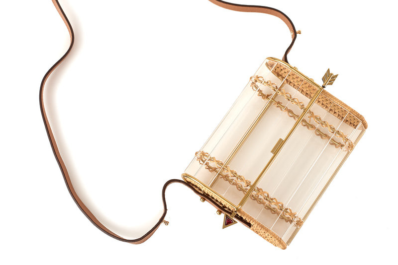 Clutch em acrílico transparente com laterais em rattan, detalhe de costura em X dourado e fecho em formato de flecha metálica dourada com ponta vermelha.