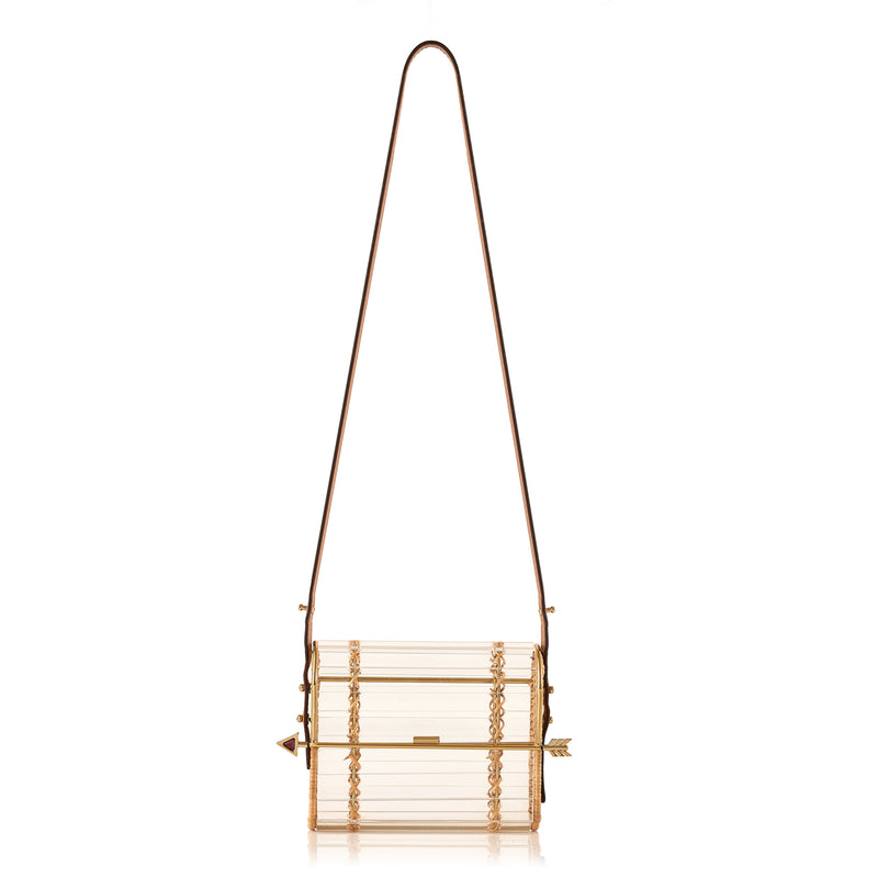 Clutch em acrílico transparente com laterais em rattan, detalhe de costura em X dourado e fecho em formato de flecha metálica dourada com ponta vermelha.